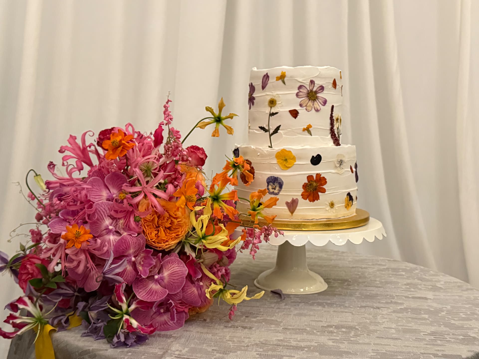 Two-tier vegan cake with pressed edible flowers beside a bright floral arrangement
